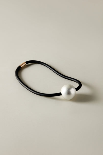 A black elastic hair tie with a large silver bead rests on a neutral background, showcasing the minimalist and contemporary design of the Single Sphere Necklace.