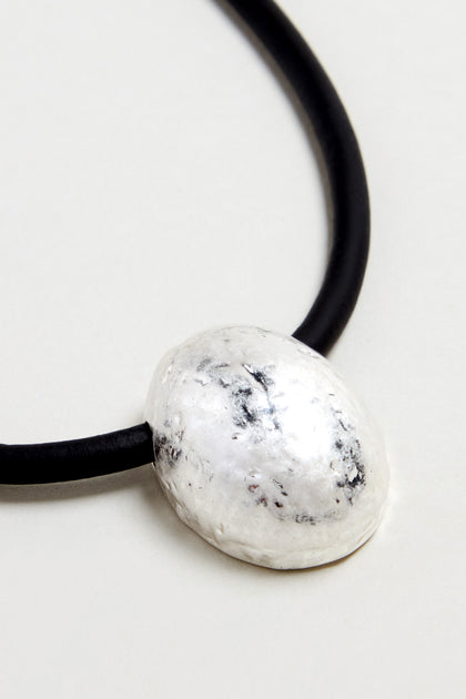 A close-up of the Hammered Pebble Necklace shows its black rubber cord and oval hammered metal pendant resting on a light surface.