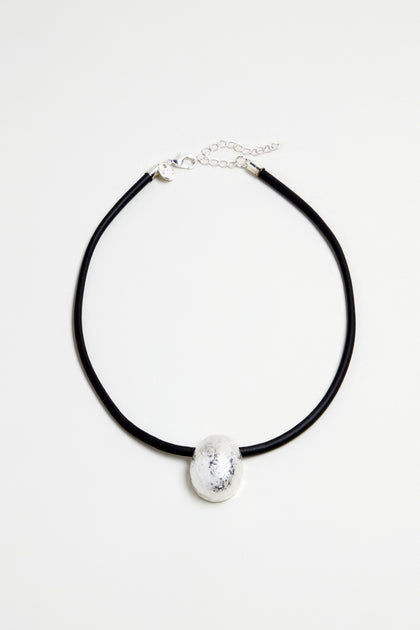 The Hammered Pebble Necklace features a black rubber cord with a large, round hammered metal pendant and clasp closure, shown on a plain white background.