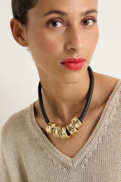 Woman with wavy dark hair wearing a blue top and the Hammered Cylinders Necklace, looking to her left against a plain background.