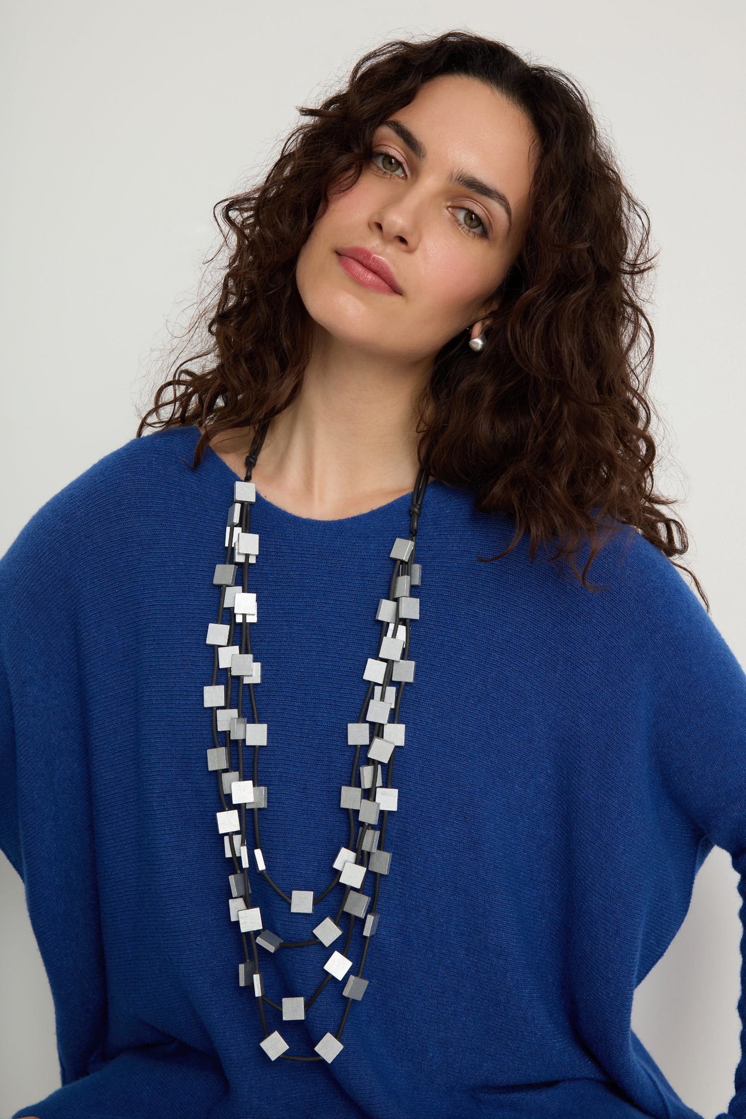 A woman with curly brown hair models the Cascade Squares Necklace—featuring silver-tone squares and rectangles—with a blue sweater, posed against a plain light background.
