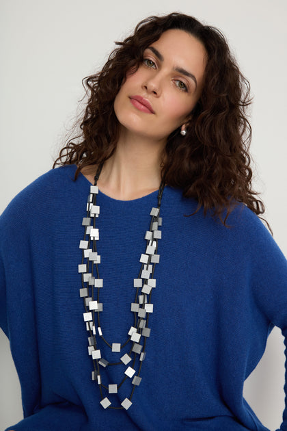 A woman with curly brown hair models the Cascade Squares Necklace—featuring silver-tone squares and rectangles—with a blue sweater, posed against a plain light background.