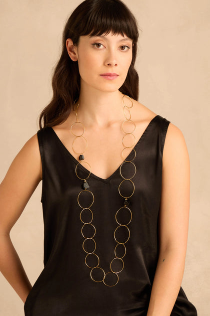 A woman wears the Midnight Stone Link Necklace, a long gold-tone hoop design featuring thin rings interspersed with small, irregular black stones.