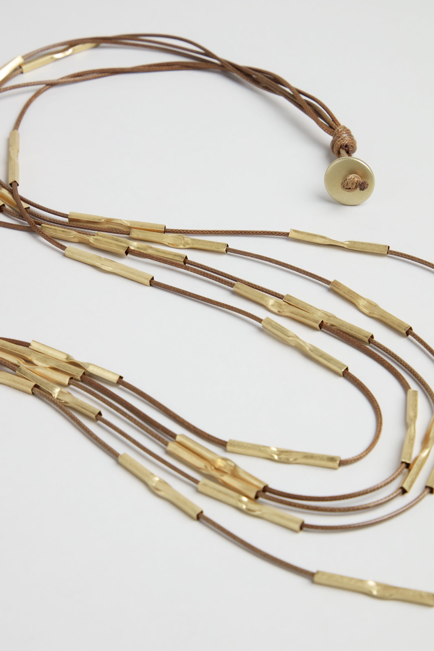The Leather and Bead Cascade Necklace features long brown leather strands, each adorned with hammered gold cylindrical beads and finished with a round gold-toned button clasp for a striking layered look.