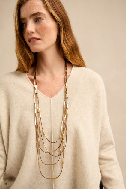 Woman with long auburn hair wearing a cream-colored V-neck sweater and multiple layered gold necklaces, including the striking Leather and Bead Cascade Necklace, looking slightly to the side against a neutral background.