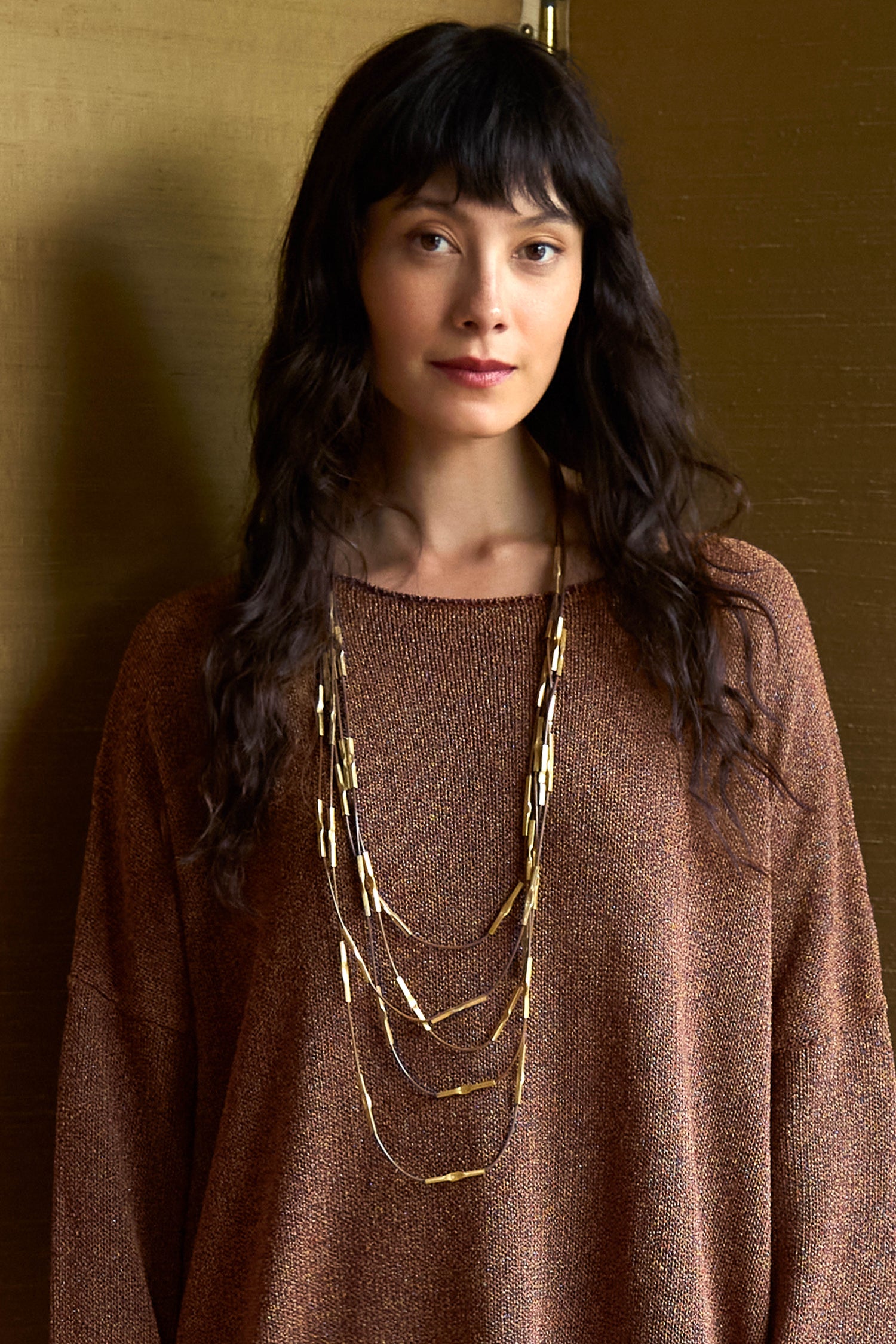 A woman wearing the Beaded Leather Cascade Necklace, a multi-strand design of dark leather cords and hammered gold metal tubes.