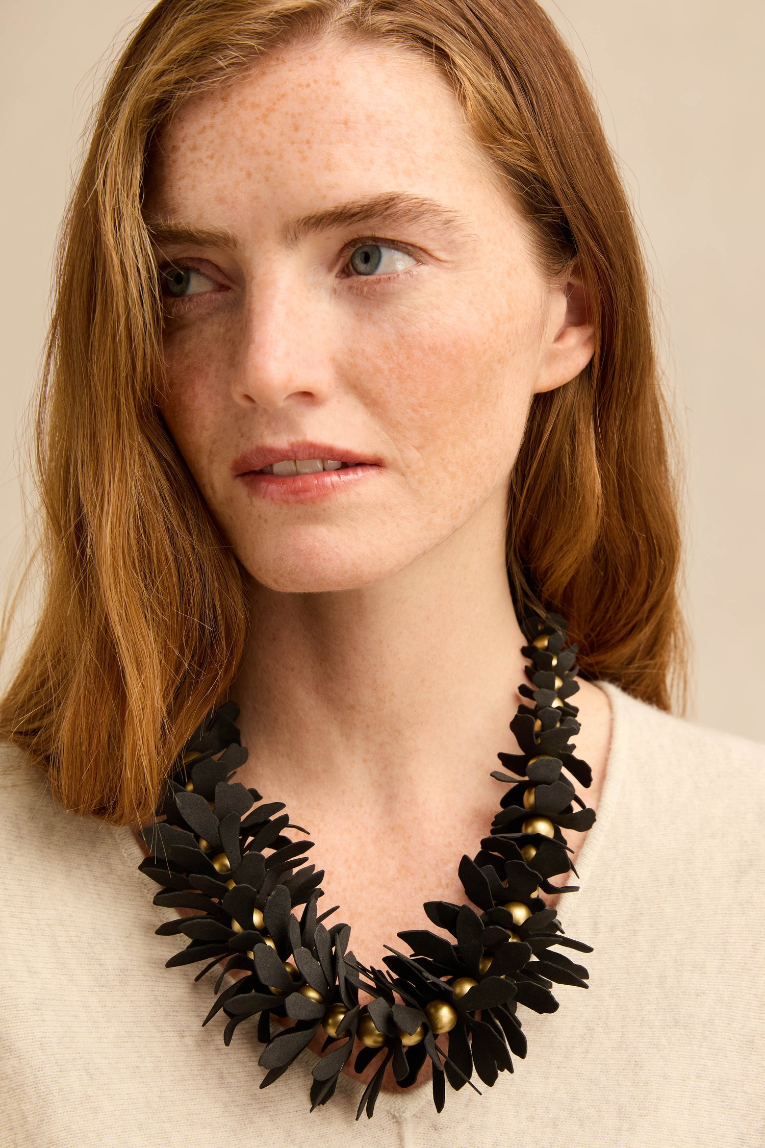 A woman with long red hair wears a cream top and the Flower Bead Necklace—a collar-length, chunky black statement necklace with gold accent beads—looking slightly to the side.