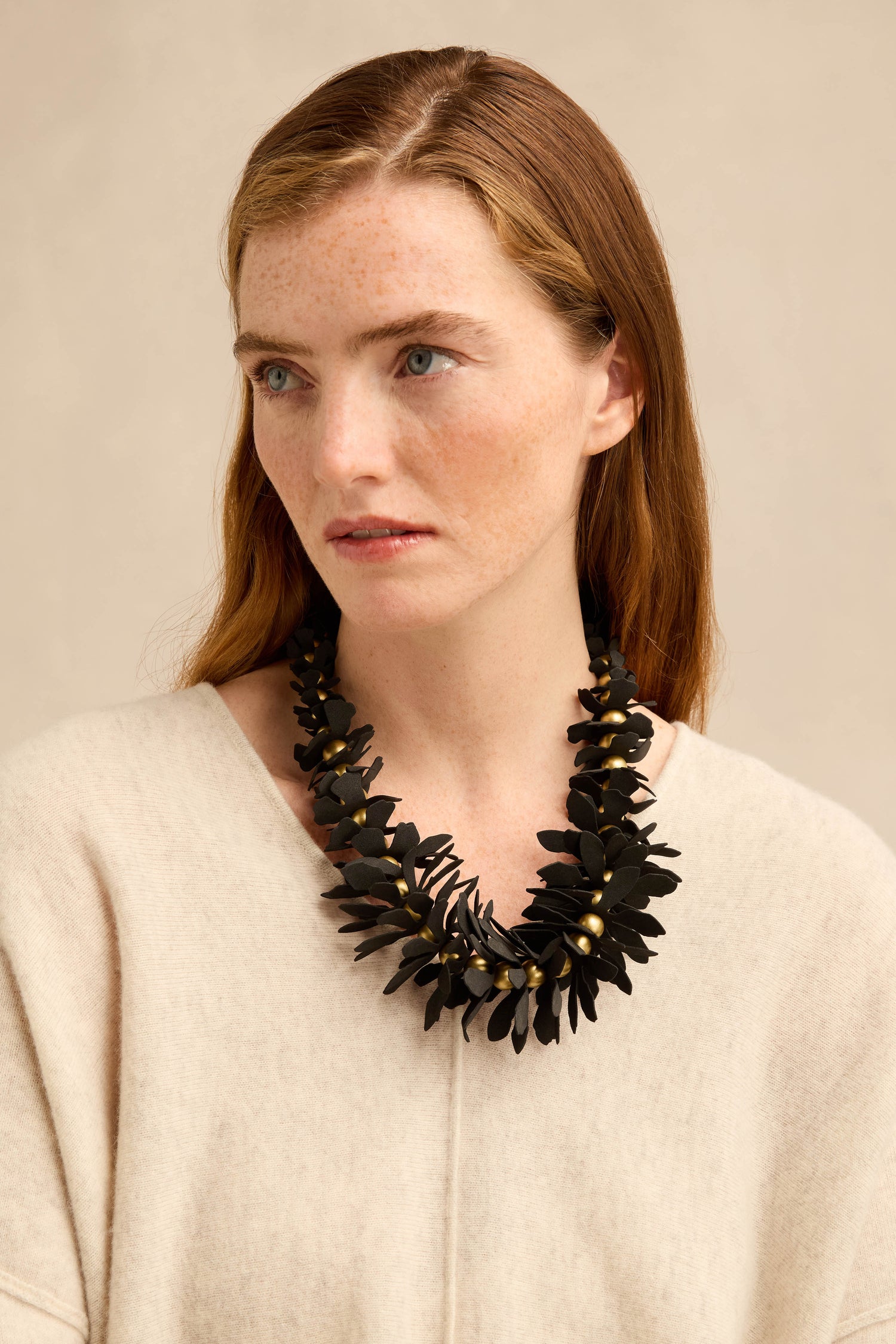 A woman with long brown hair wears a cream sweater and the Flower Bead Necklace, which features collar-length flower beads with gold accents, as she looks slightly to the side against a neutral background.