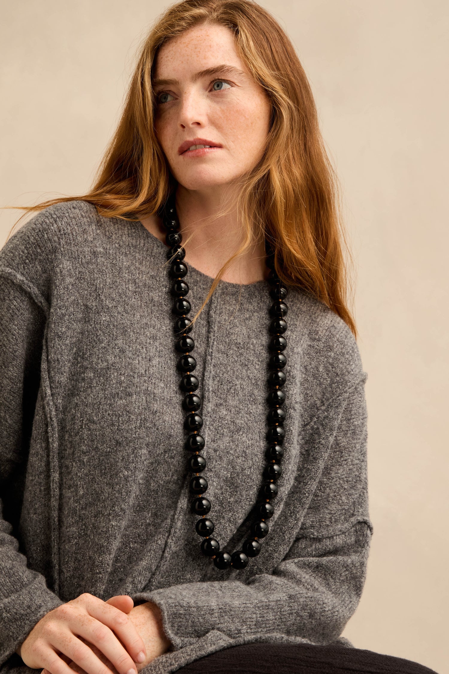 A woman with long auburn hair wears a loose grey sweater and the Long Spheres Necklace, featuring stylish resin spheres, while sitting against a plain beige background.