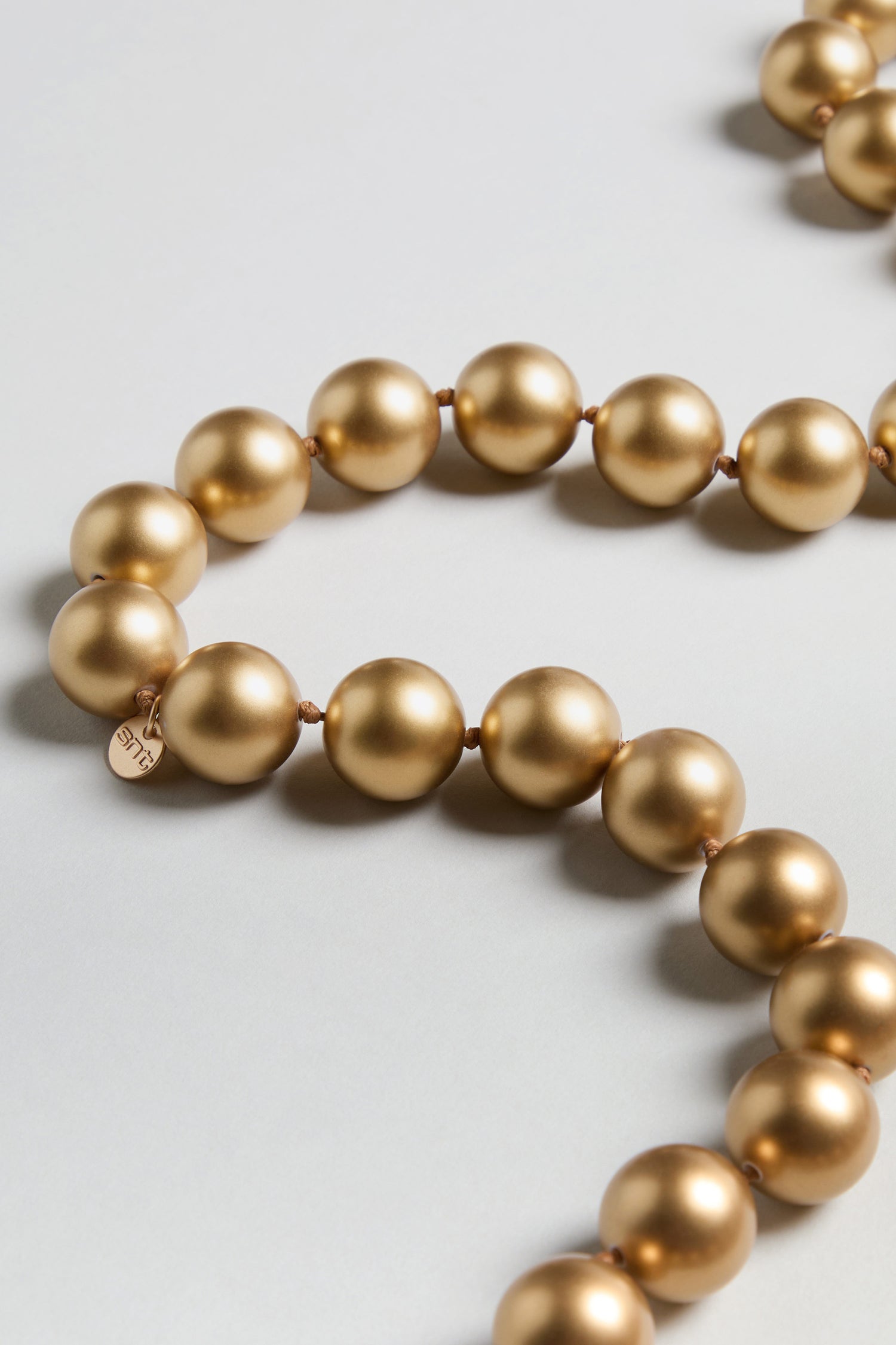 A close-up of the Long Spheres Necklace, showcasing resin spherical beads with a gold burnished finish and a small round tag, elegantly arranged on a light-colored surface.