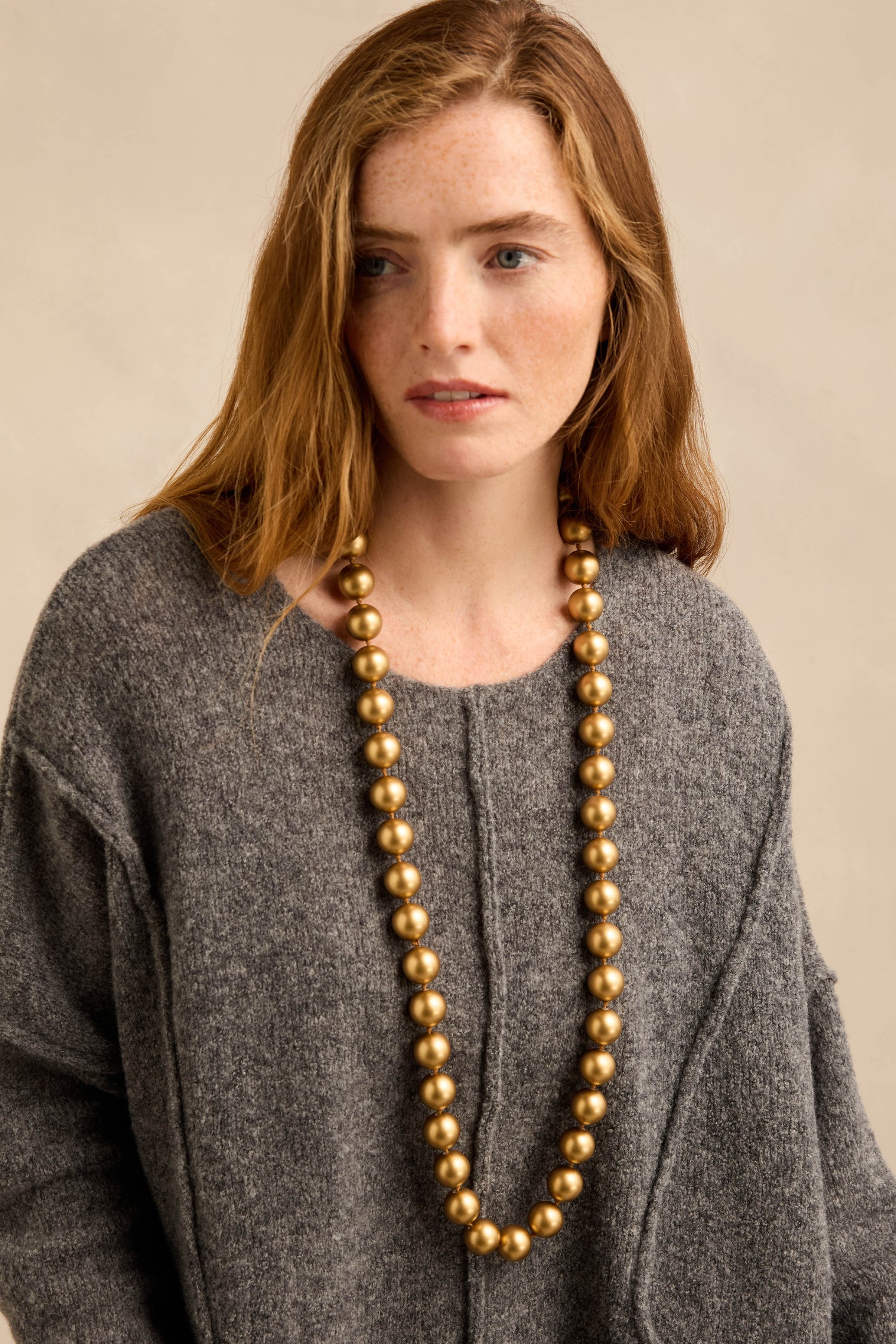 A woman stands against a beige background, wearing a grey sweater and the Long Spheres Necklace—a longline gold beaded piece with a subtle burnished finish.