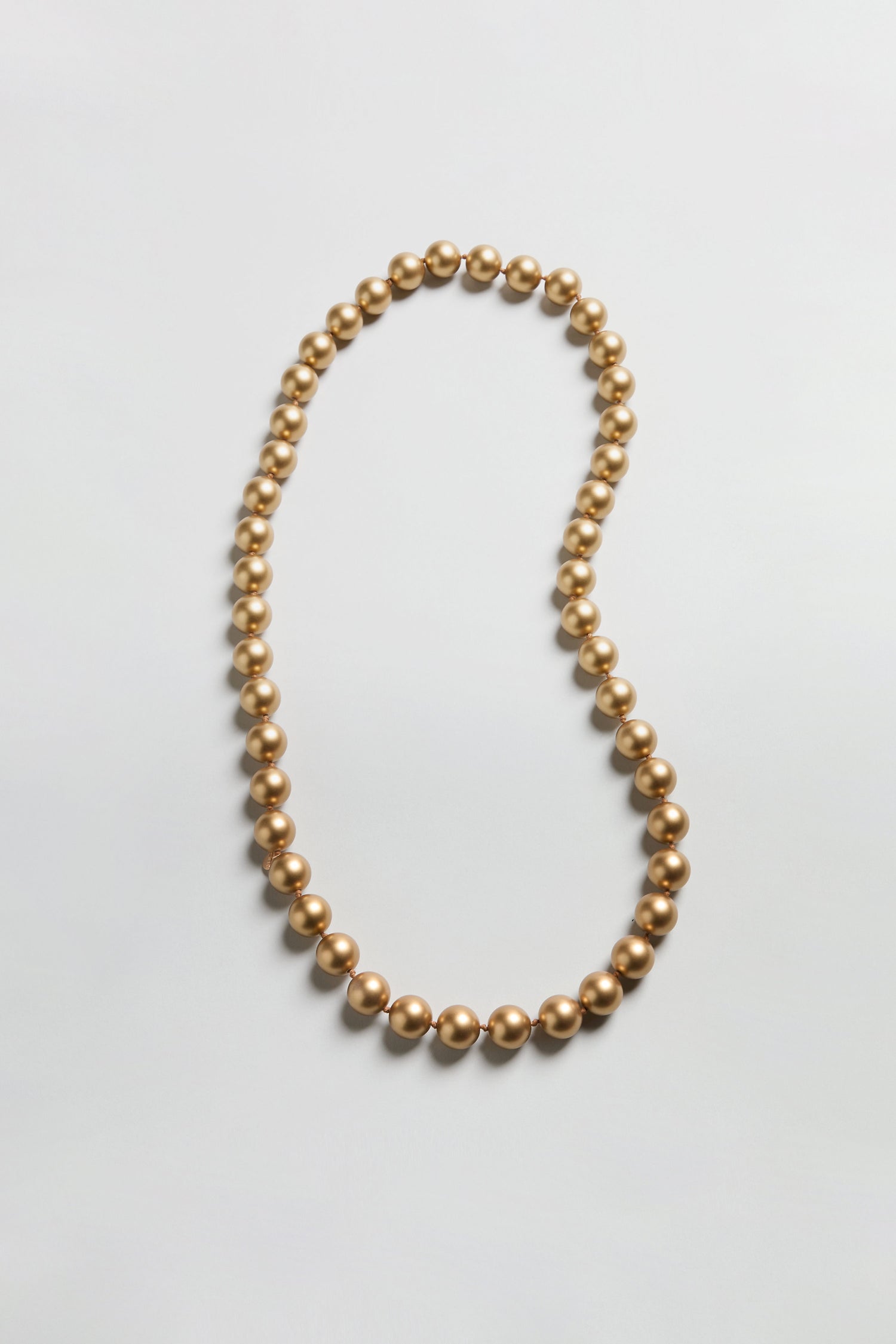 The Long Spheres Necklace is a single strand longline piece with round, gold-colored beads and a stunning gold burnished finish, displayed in an open loop on a simple, light background.