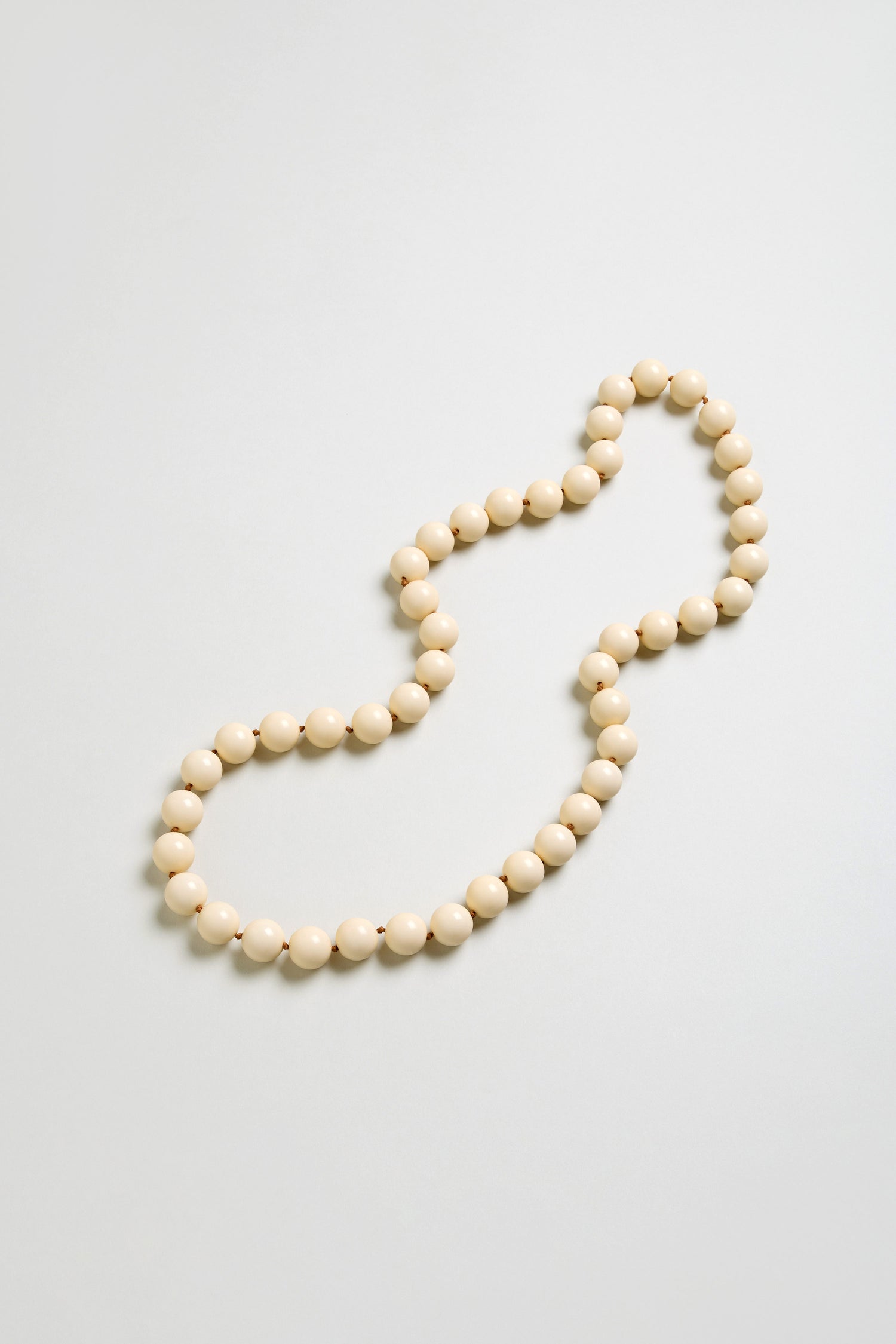 A single strand of round, cream resin beads forms a loose, irregular loop on a light grey background, highlighting the elegant simplicity of the Long Spheres Necklace.