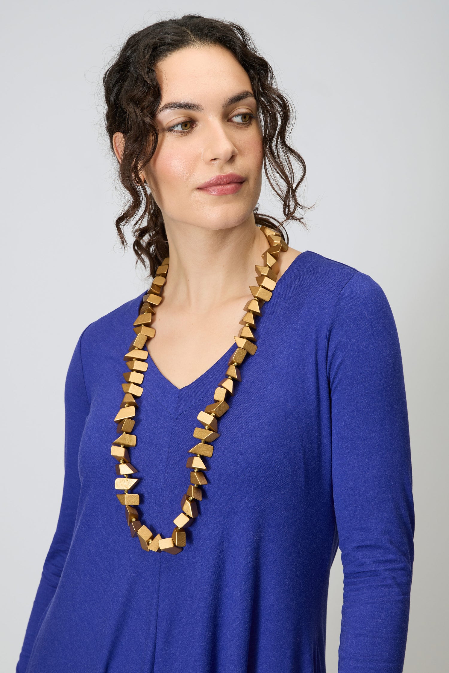 A woman showcases the Long Wooden Metallic Necklace, featuring metallic-coated wooden beads, paired with a blue V-neck top as she looks slightly sideways against a plain background.