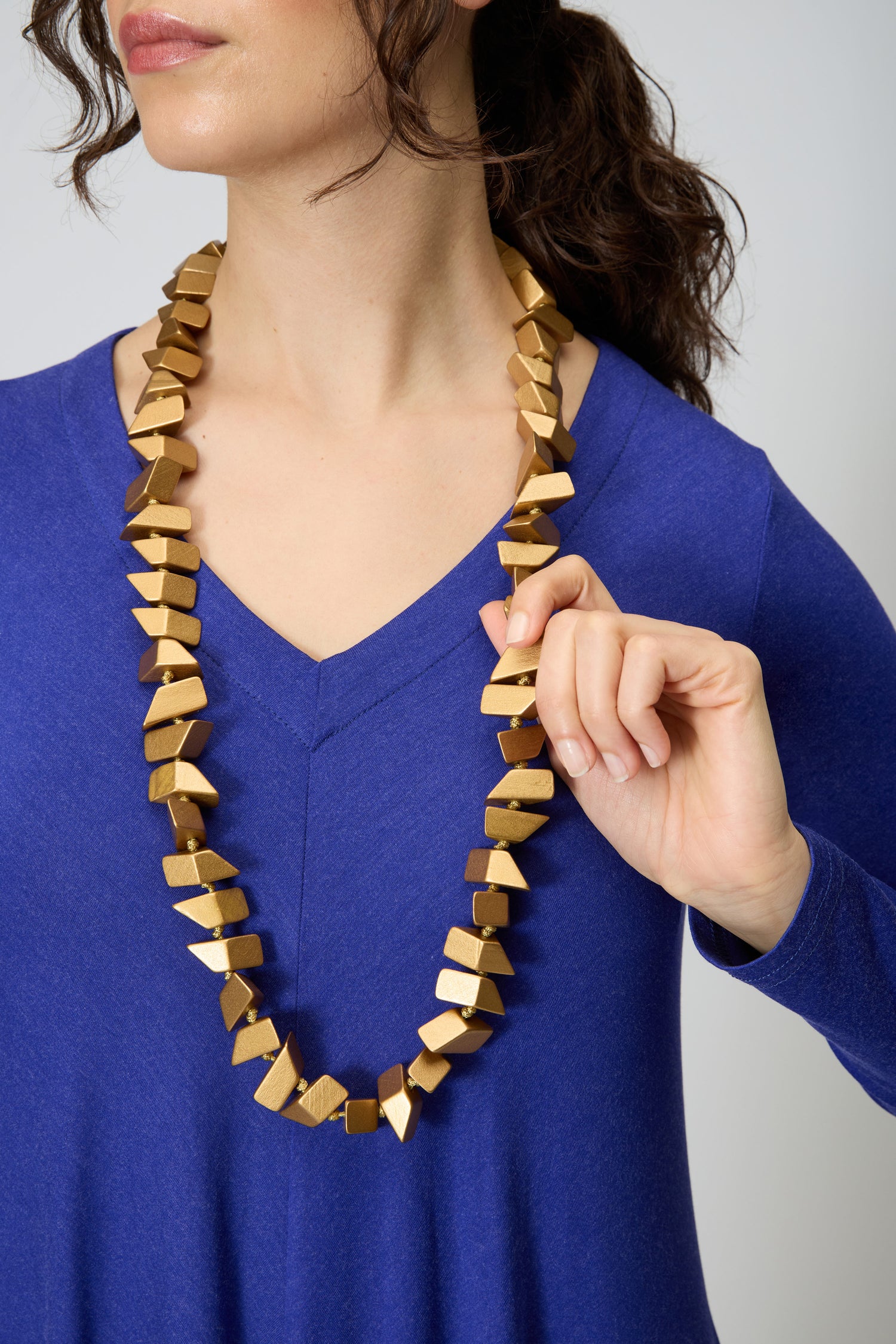 A woman wearing a blue v-neck top holds the Long Wooden Metallic Necklace—a lightweight statement piece featuring metallic-coated wooden beads and a gold geometric design.