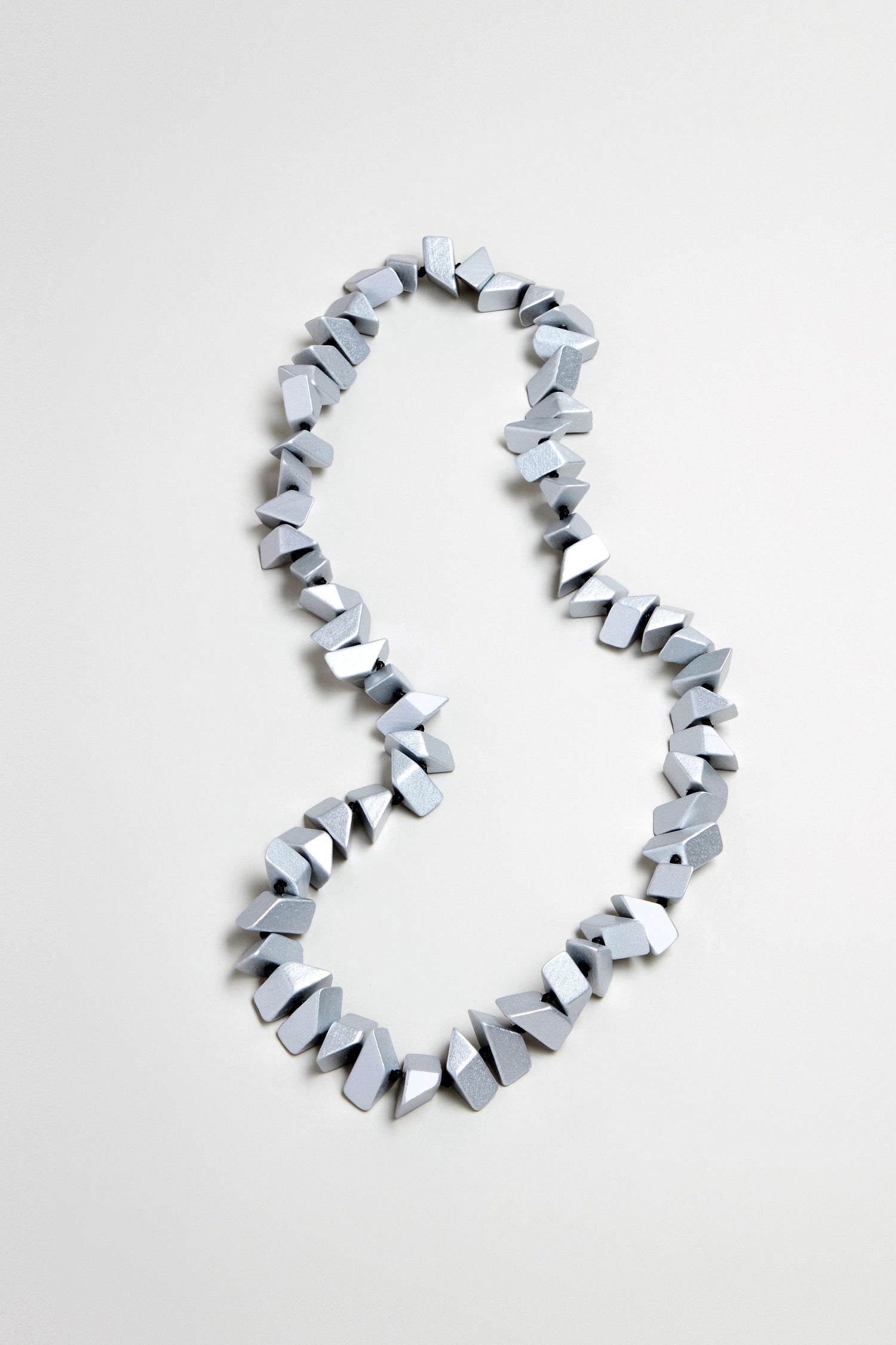 The Long Wooden Metallic Necklace, featuring irregularly shaped, silver-gray faceted wood beads, is displayed on a plain light surface.