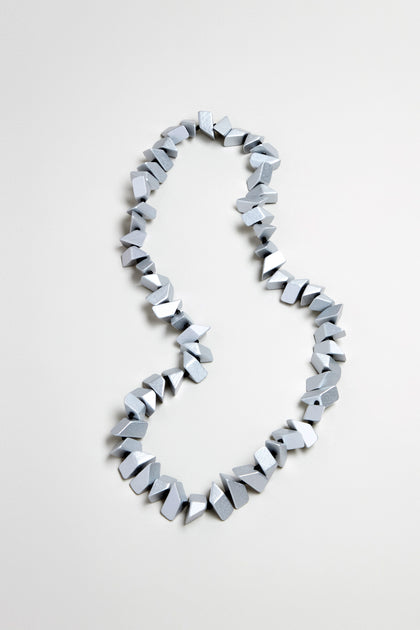 The Long Wooden Metallic Necklace, featuring irregularly shaped, silver-gray faceted wood beads, is displayed on a plain light surface.