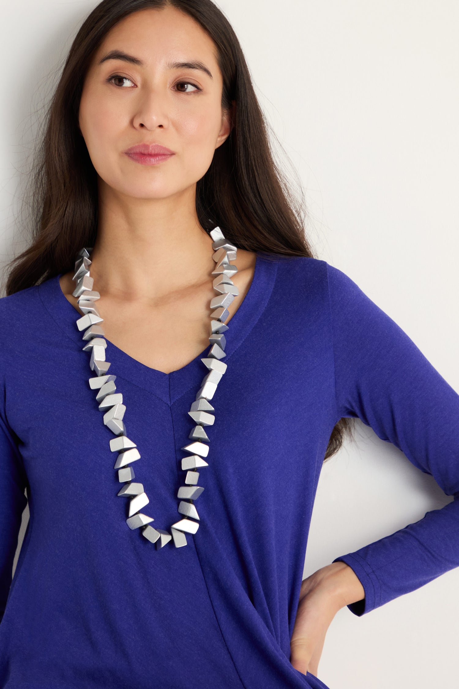 A woman with long dark hair wears a cobalt blue long-sleeve top and the Long Wooden Metallic Necklace, featuring faceted wood beads, as she looks slightly to the side against a plain background.