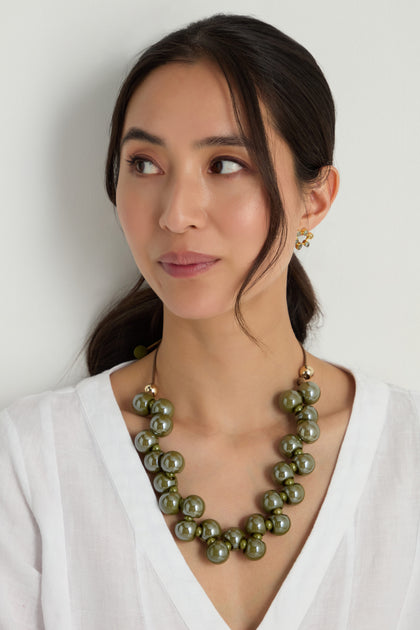 Woman with long dark hair in a white blouse wears the Macrame Resin Bead Necklace—chunky green resin beads on an adjustable cord—and small gold earrings, looking slightly to the side against a plain background.