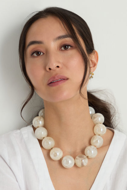 A woman with long dark hair wears a white blouse, the Marble Spheres Necklace featuring a marbled finish, and small gold earrings, posing against a plain light background.