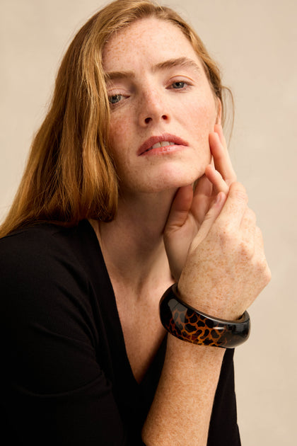 A woman with long red hair, dressed in a black top, poses with her hand near her face to showcase the Leopard Bangle—a wide resin accessory featuring a glossy leopard print finish.