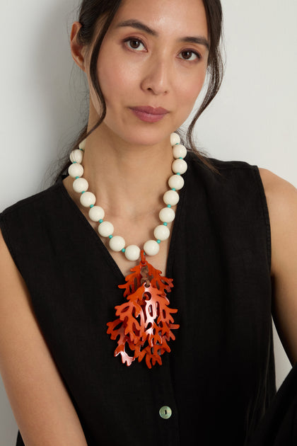 A woman in a black sleeveless top wears the Coral Resin Short Pendant Necklace, featuring chunky resin beads and a bold orange coral pendant, seated against a plain background.