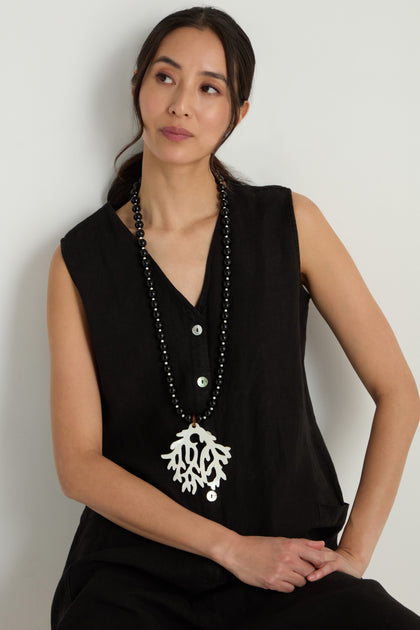 Woman wearing a sleeveless black top and the Coral Resin Pendant Necklace, a sculptural long drop necklace with a large white coral resin pendant, sits against a light background.
