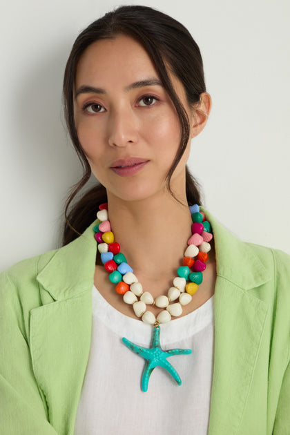 A woman stands against a plain light background, wearing a light green blazer, white top, and the Turquoise Starfish Pendant Necklace with colorful resin beads and a large turquoise starfish charm.