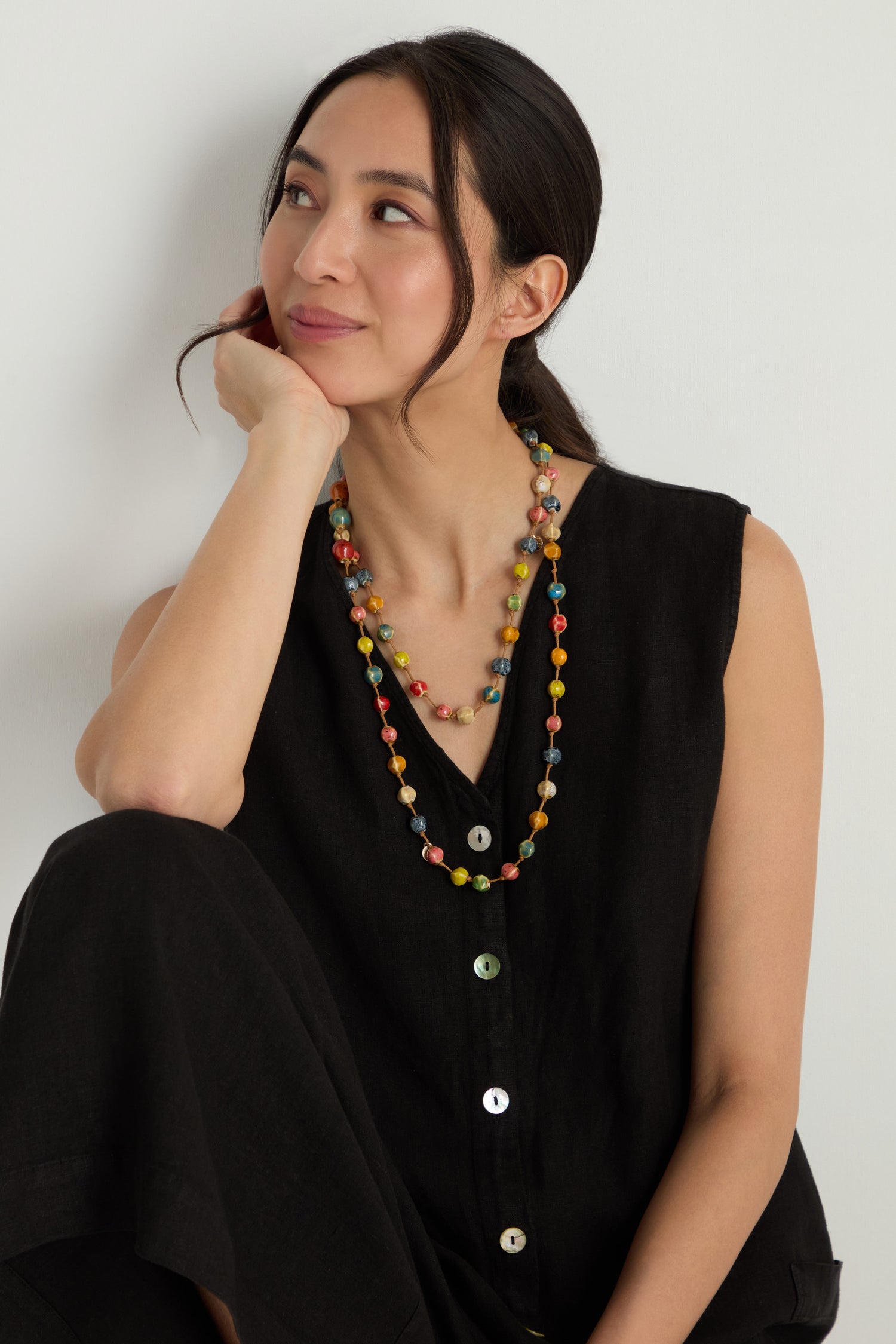 A woman in a sleeveless black buttoned dress sits with her hand on her cheek, wearing the Rainbow Ceramic Bead Necklace and gazing to the side.