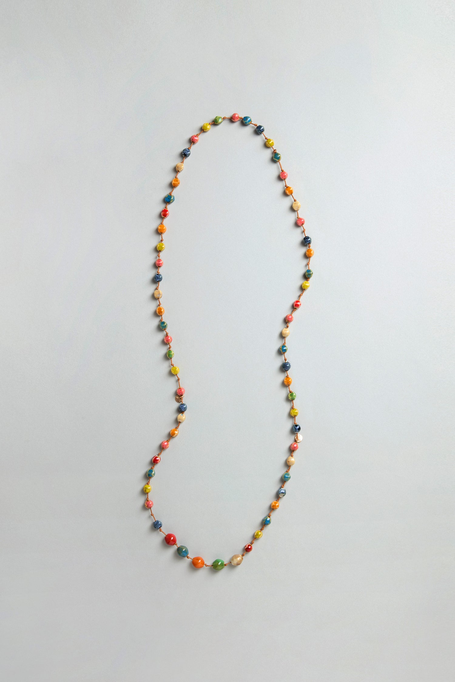 The Rainbow Ceramic Bead Necklace, featuring small multicolored ceramic beads, is displayed in a loose loop on a plain light gray surface.