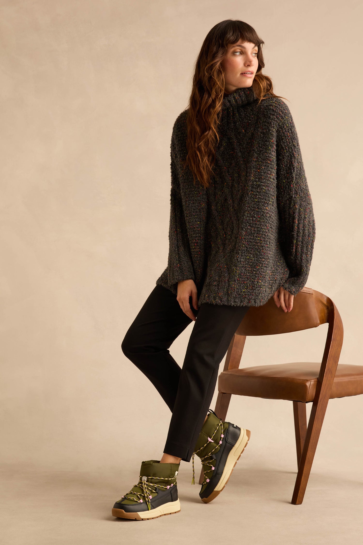 A person sitting on a wooden chair wearing dark trousers and olive green Whitney Quilted Toggle Slip on Boots.