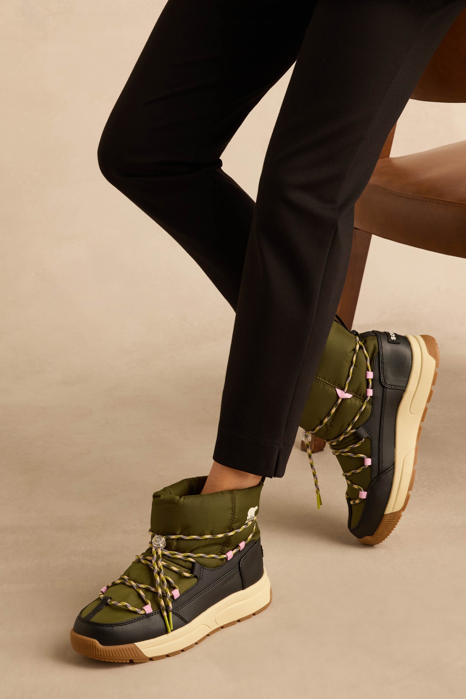Whitney Quilted Toggle Slip on Boots in olive green and black, featuring quilted insulation, yellow and pink laces, and worn with black pants.