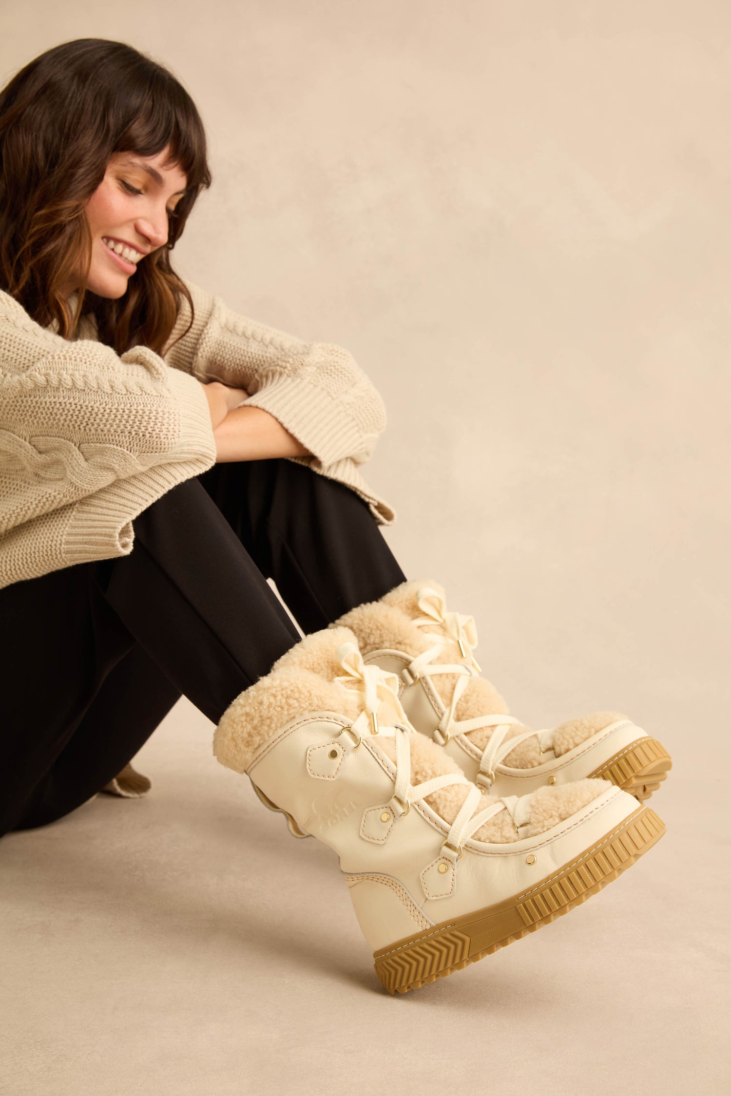 A smiling woman sits on the floor wearing cream-colored, waterproof Long Leather Alpine Boots with genuine shearling lining and a thick rubber sole.