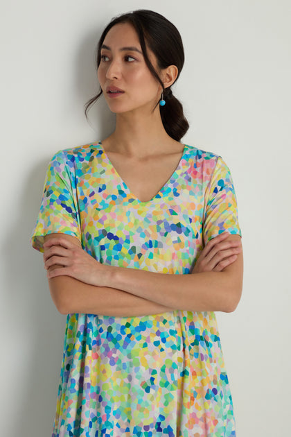 Woman with dark hair in a low ponytail, wearing the Summer Confetti Jersey Bubble Dress—short sleeves, V-neck, colorful pattern—stands against a plain light wall with arms crossed, looking to the side.