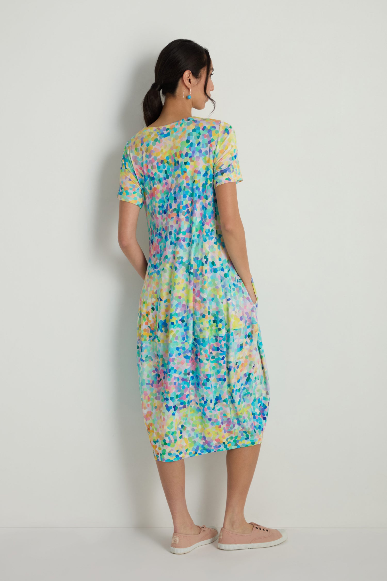 A woman faces away from the camera, standing against a white wall in the Summer Confetti Jersey Bubble Dress—a short-sleeve, knee-length style with a colorful abstract print and relaxed fit—paired with light sneakers for an easy, effortless look.