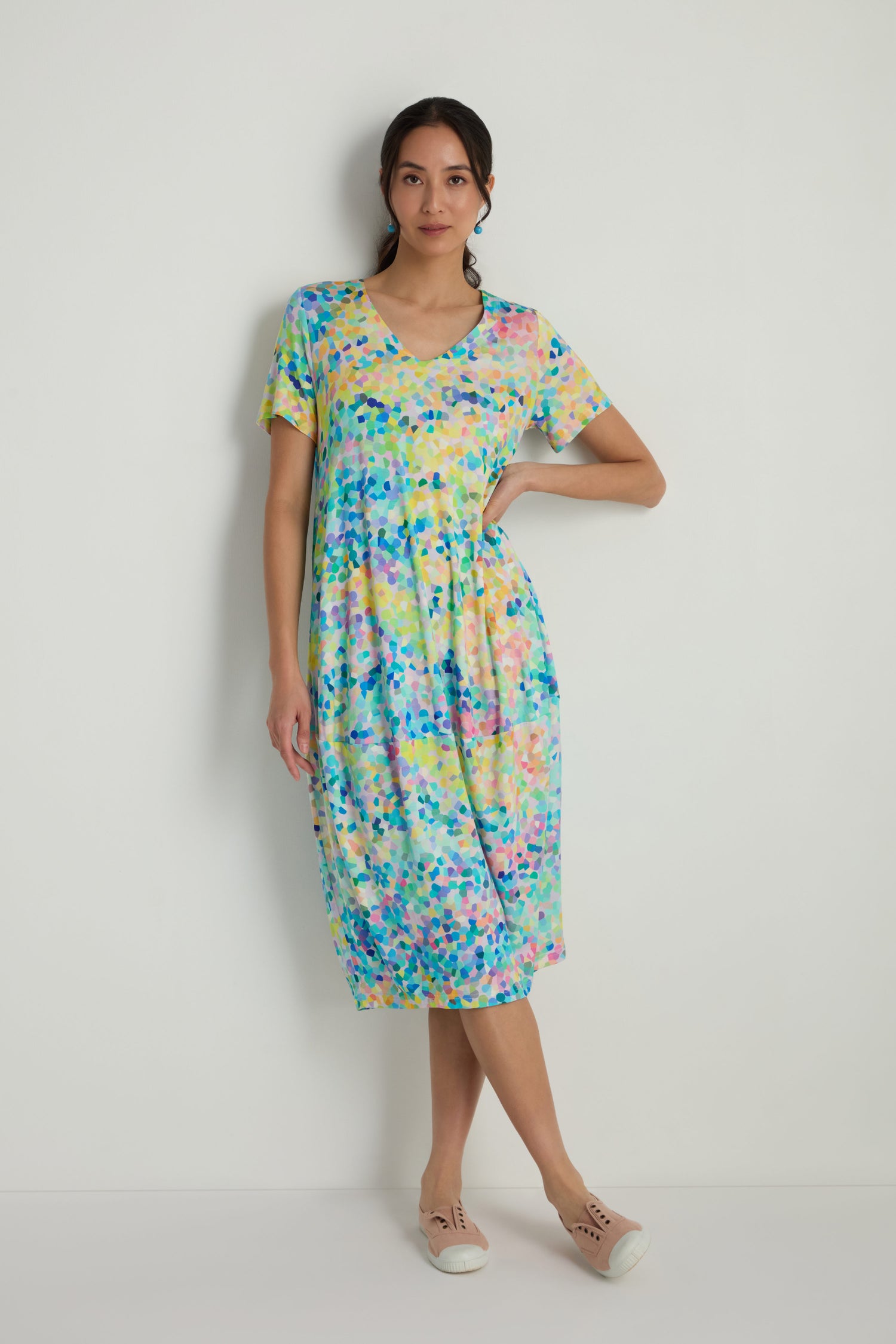 A woman stands against a plain white wall wearing the Summer Confetti Jersey Bubble Dress with a relaxed fit, short sleeves, V-neck, abstract pattern, knee length, and beige slip-on shoes.