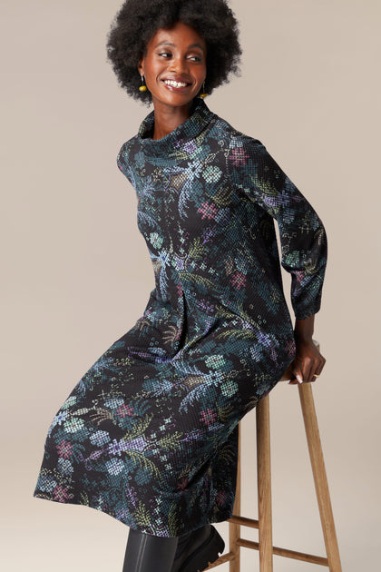 A woman is sitting on a stool in a Pixelated Tapestry Jersey Dress, adorned with deep hues of black floral patterns.