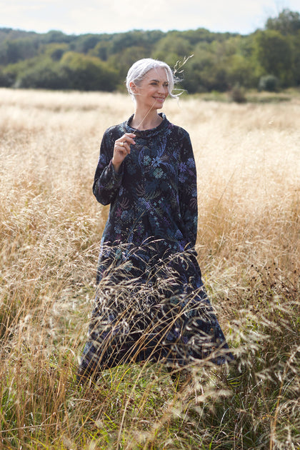 An older woman wearing a Pixelated Tapestry Jersey Dress stands majestically in a field of tall grass, creating a mesmerizing fusion of hero print and pixelated tapestry.
