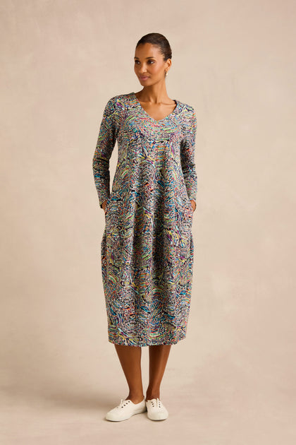 A woman stands against a neutral background wearing the colorful Patchwork Fields Jersey Bubble Dress, featuring long sleeves, convenient pockets, and white sneakers.
