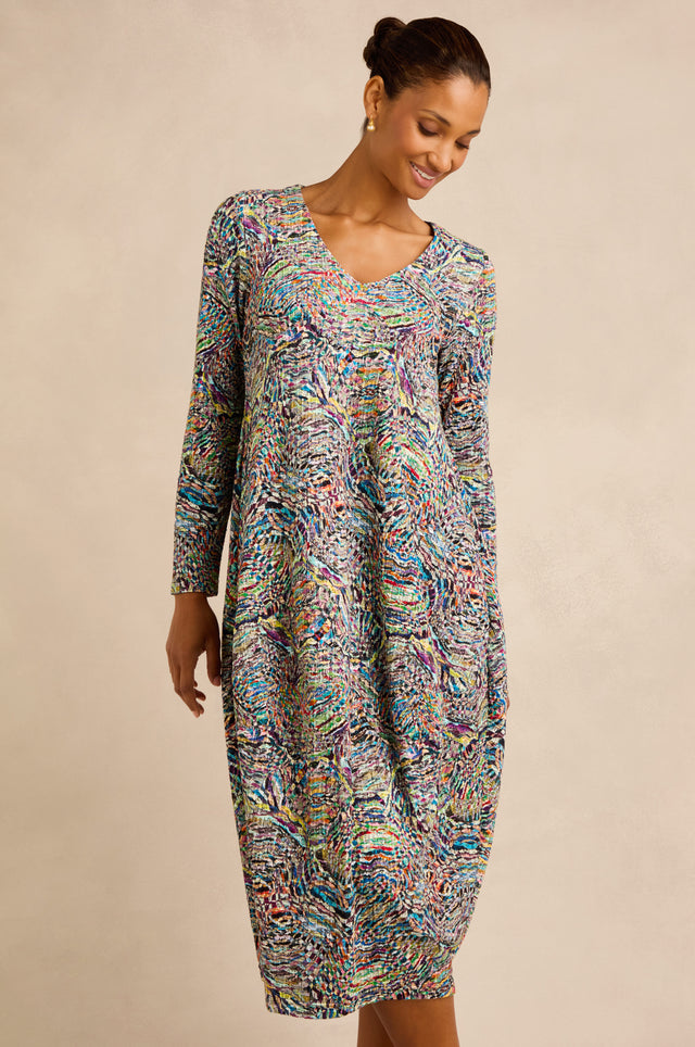 A woman wearing the Patchwork Fields Jersey Bubble Dress—a long-sleeved, multicolored patterned midi dress with a bubble silhouette—stands against a neutral background and looks down with a slight smile.