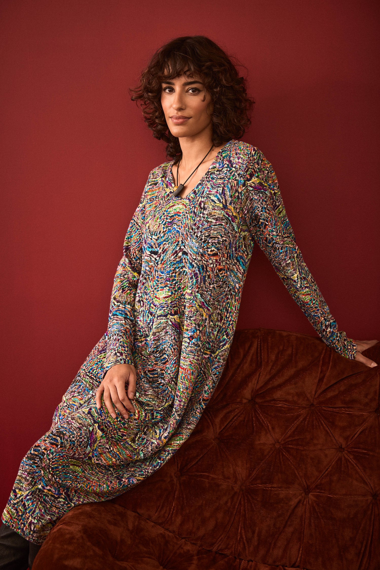 A person with curly hair wearing the Textured Prism Jersey Bubble Dress sits on a brown cushioned couch against a dark red wall.