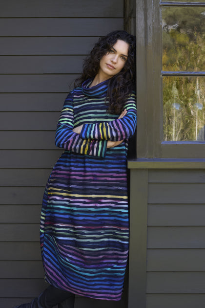 Person in a Textured Watercolour Landscape Cowl Dress leans against the corner of a dark-colored wooden house with crossed arms.