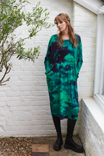 A woman in a green Jewelled Sky Jersey Dress made from Lenzing EcoVero™ Viscose™ fabric, standing in front of a brick wall.
