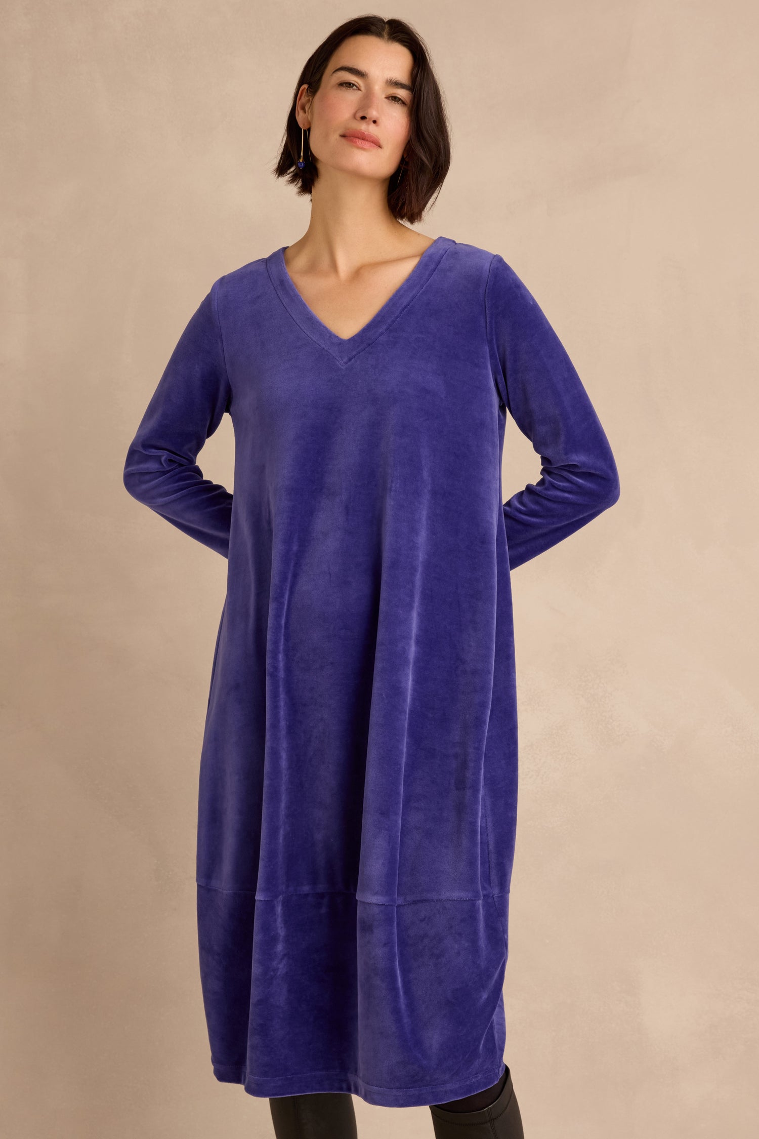 A woman wearing the Velvet Jersey Bubble Dress—a loose-fitting, long-sleeve, deep blue cotton-rich velvet jersey dress with a V-neck—stands against a plain beige background.