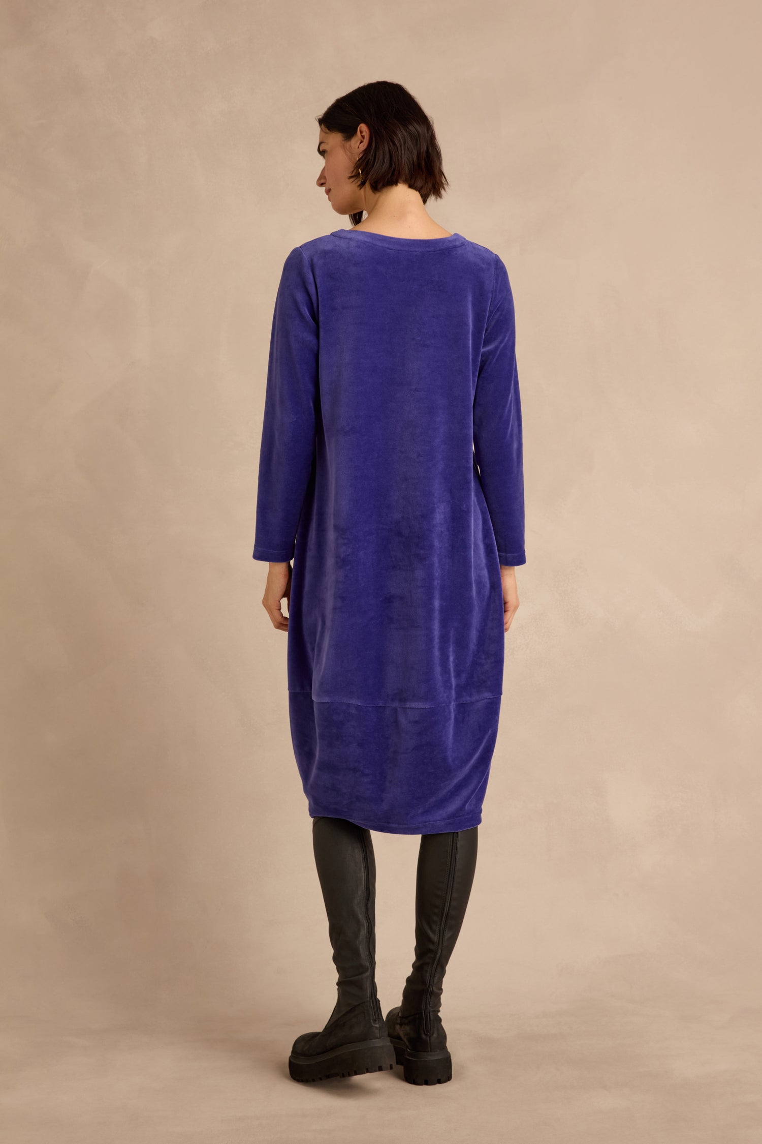 A person stands with their back to the camera, wearing the Velvet Jersey Bubble Dress—a long-sleeved, knee-length cotton-rich velvet dress in purple—paired with black leggings and black shoes, set against a beige background.