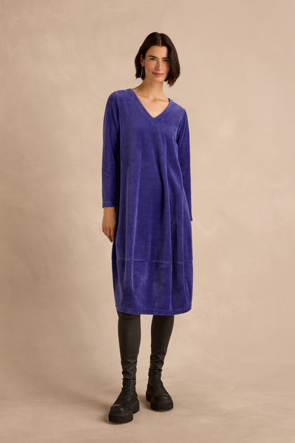 A person stands against a beige background wearing the Velvet Jersey Bubble Dress in purple, paired with black leggings and chunky black boots.