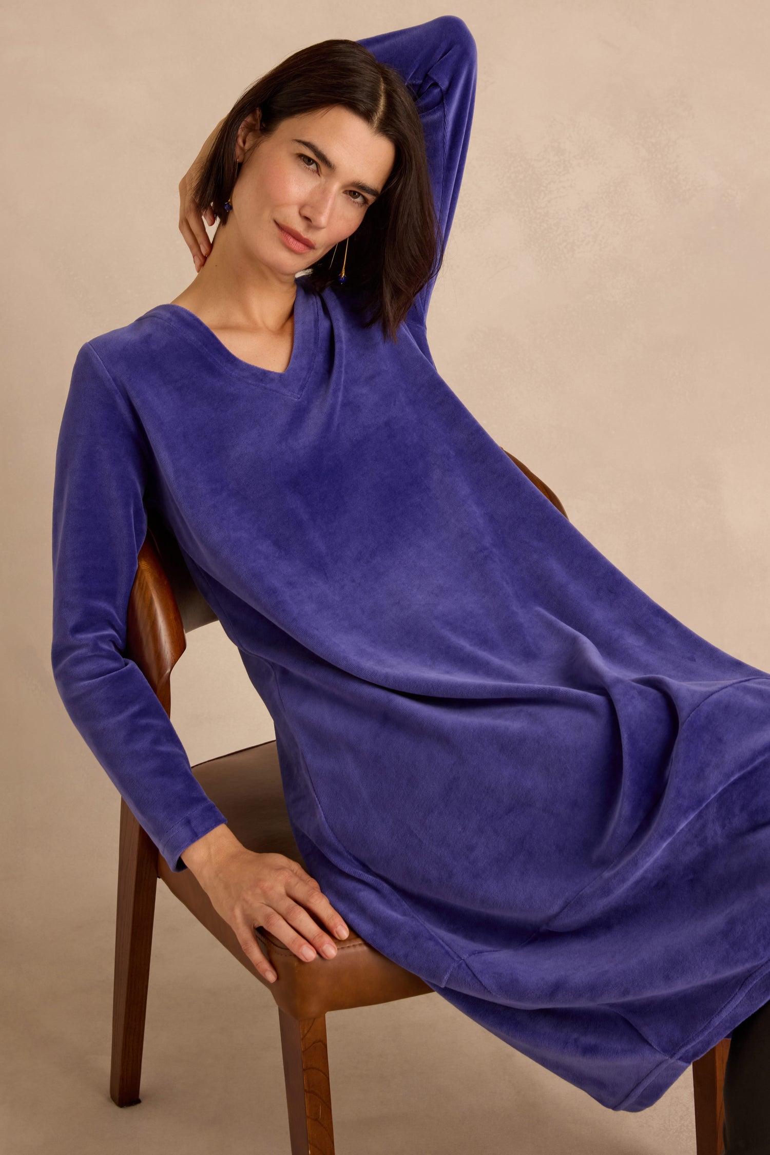 A woman in a Velvet Jersey Bubble Dress with long sleeves and a purple hue sits on a wooden chair, one arm draped over the chair back and the other resting on her lap, set against a neutral background.