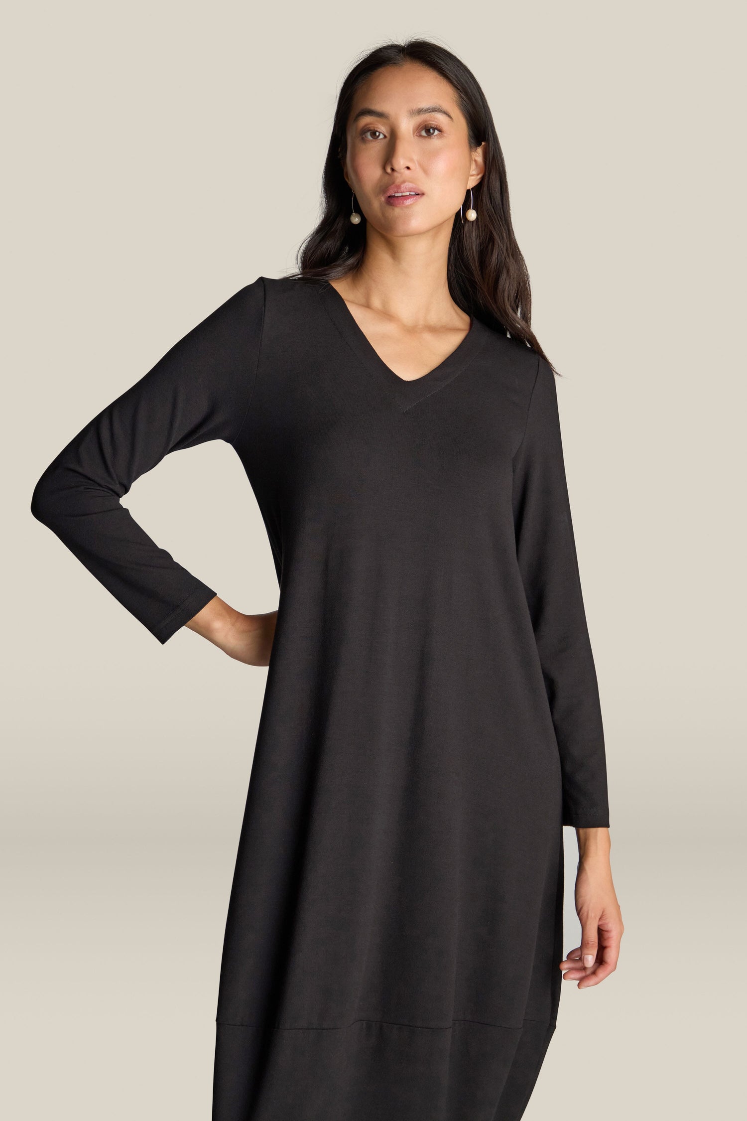 A person stands wearing the Fluid Crepe V Neck Bubble Dress, which features long sleeves in a black fluid crepe material and showcases a universally flattering silhouette.