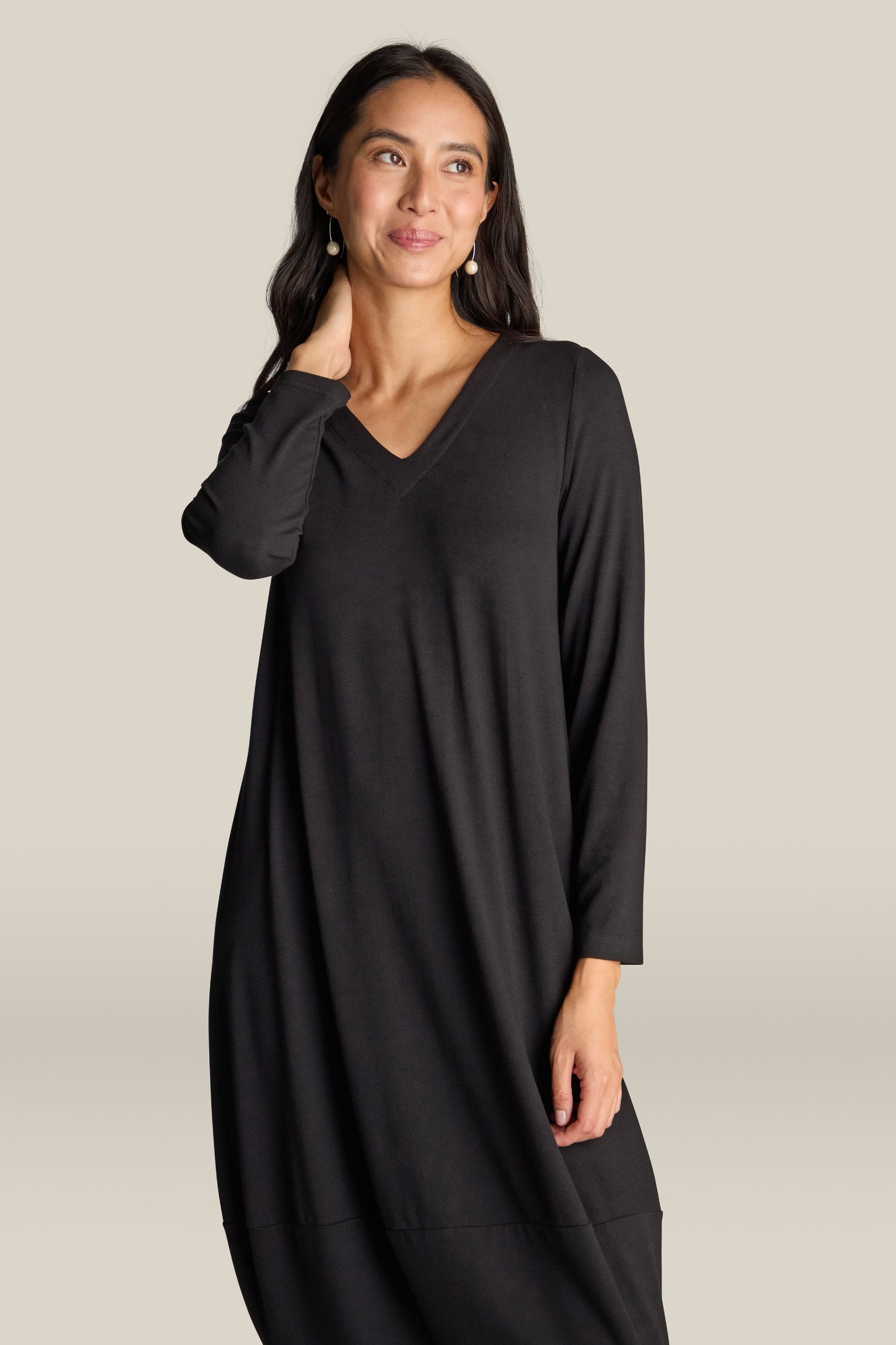 A person with long hair, dressed in a Fluid Crepe V Neck Bubble Dress, stands against a neutral background. They have one hand touching their neck and wear a slight smile on their face.