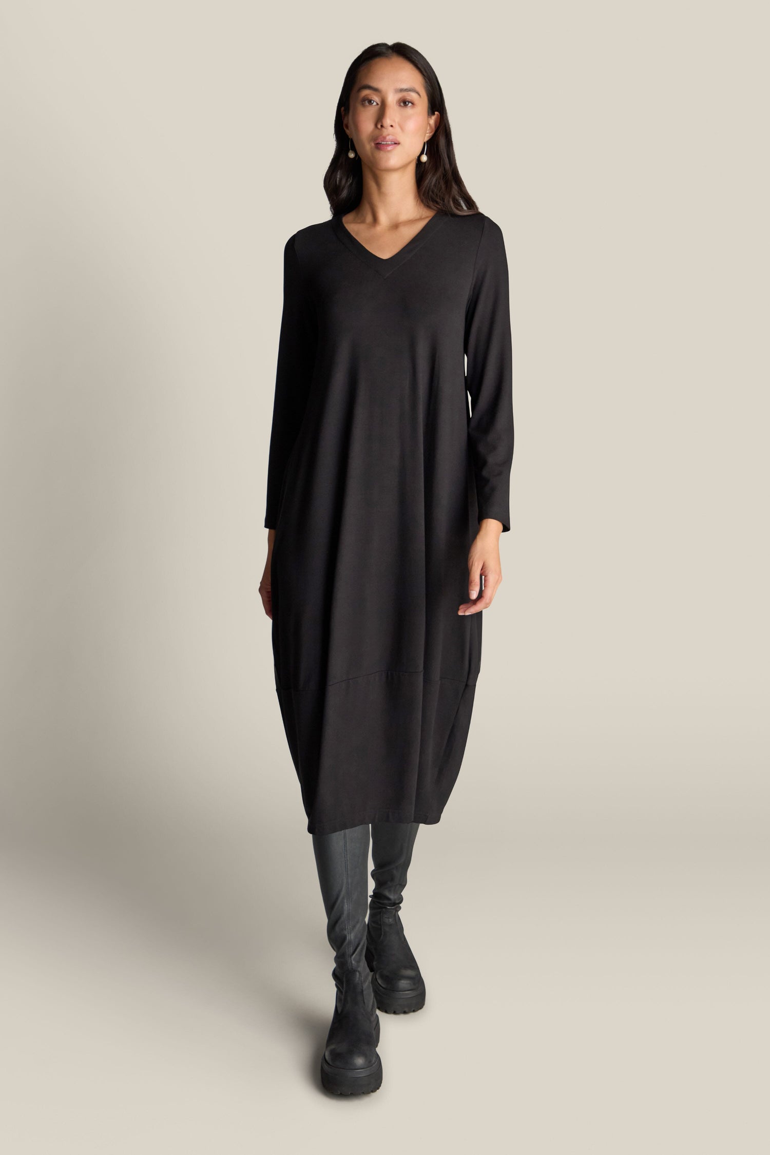 A person stands against a plain background wearing the Fluid Crepe V Neck Bubble Dress, a black, long-sleeved, midi dress crafted from fluid crepe material. It is paired with dark pants and black boots. This universally flattering silhouette offers timeless elegance.